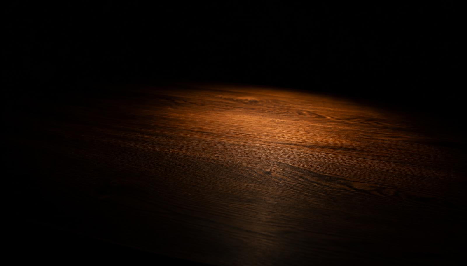 An empty dark desk surface with a single warm pool of light falling across the grain